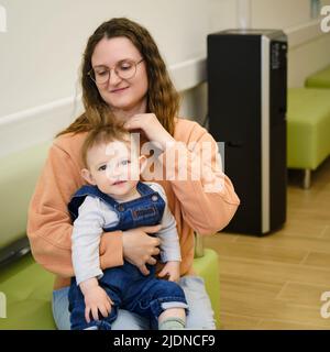 A mother woman with a toddler baby on a couch in the corridor of the clinic. Mom and child in the lobby of the polyclinic. Kid aged one year Stock Photo