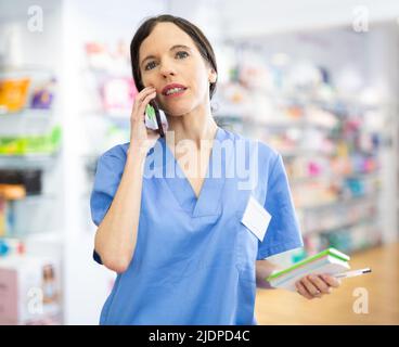 Female pharmacist talking on mobile phone and taking notes in a notebook in pharmacy Stock Photo