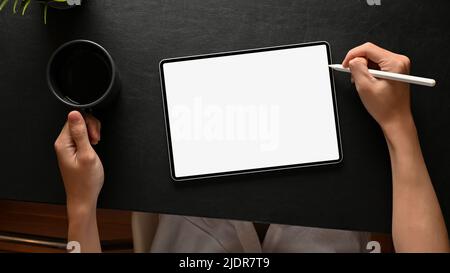 Top view, A man graphic designer designing his artwork on digital tablet touchpad with stylus pen and sipping morning coffee at his modern dark office Stock Photo