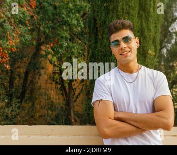 A guy with sunglasses smiling slightly resting on low wall with his arms crossed. Stock Photo
