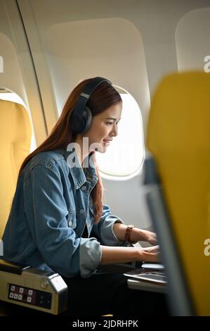 Beautiful asian female passenger in the economy class sits at the window seat listening music on headphones and using portable laptop computer. Plane Stock Photo
