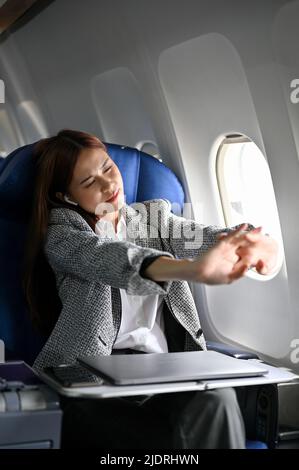Beautiful asian young businesswoman stretching arms, feeling uncomfortable and tired during a long flight flying to another country. Business trip con Stock Photo