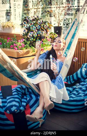 A young woman is sitting in a summer outdoor cafe in hammock and blowing soap bubbles. Stock Photo