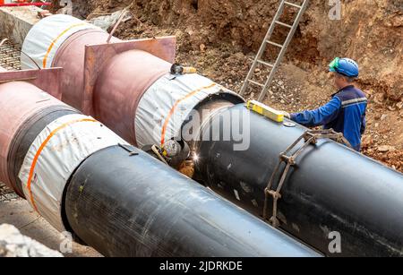 Samara, Russia - June 18, 2022: Worker with protective mask welding metal with sparks. Construction works on large iron pipes at a depth of excavated Stock Photo