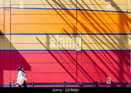 Lady in a white coat walking in front of a colorful wall. Shadow of a metallic structure. Brussels. Stock Photo