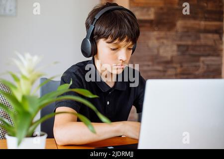 Portrait of concentrated teenager boy wearing black T-shirt, headphones sitting at desk, using laptop, surfing internet. Stock Photo