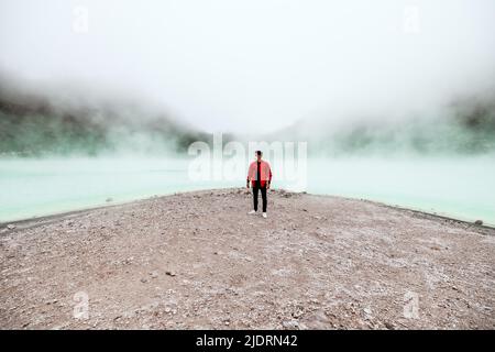 wide angle young man in red bomber jacket standing at kawah putih sulfer lake in Bandung Stock Photo