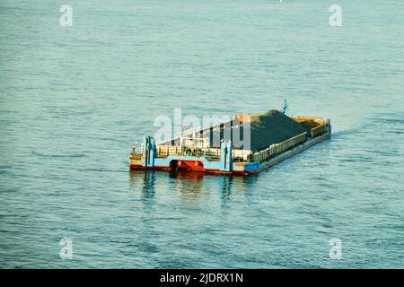 A river non-propelled barge barge (side stone dump vessel) with rubble is anchored in the middle of the river waiting for a tug Stock Photo