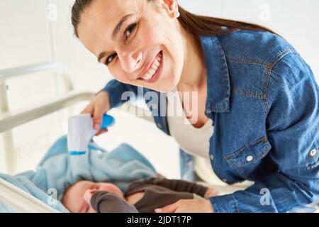 Smiling mother measuring baby's fever in bed with forehead thermometer Stock Photo