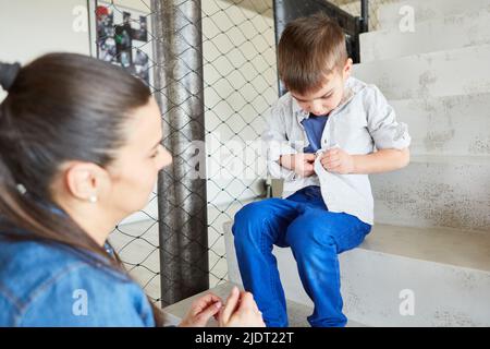 Little boy is sitting on the stairs and buttoning his shirt with mother's support Stock Photo