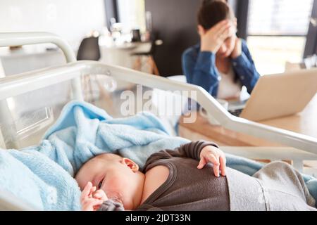 Sleeping baby in cot and exhausted single mother at pc in background Stock Photo