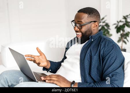 Excited happy 30s African guy enjoying virtual meeting, cheerful glad black man making video call on the laptop, sitting on the sofa at home, stay in touch with friends or family on the distance Stock Photo