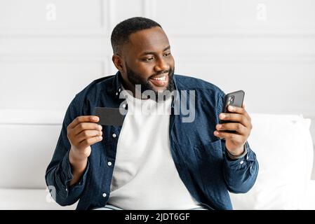Serene young guy African-American holding debit card and smartphone, sitting on the sofa at home, making online purchases, transferring money, smiling black man enjoying online shopping from home Stock Photo