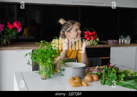Smiling, laughing blond woman in apron smelling bunch fresh juicy radish. Summer holiday at home. Farming organic food