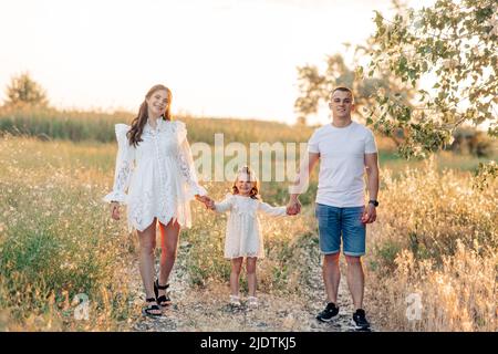 Happy parents hold hands of their daughter and walk among meadow at sunset. Stock Photo
