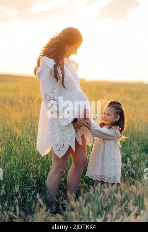 Little child girl touches belly of her pregnant mother among meadow at sunset. Stock Photo