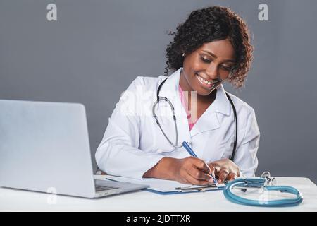 African woman doctor in a white coat and sits at a table in a medical office and works on a computer, telemedicine, online patient consultation Stock Photo