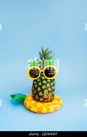 Funny pineapple in a sunglass of pineapple shape on the yellow inflatable ring on the blue vertical background. Tropical style summer on beach vibes. Stock Photo