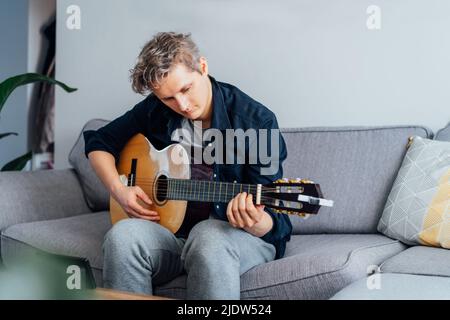 Man taking online guitar lessons via a mobile application on his phone at home sitting on the gray couch, sofa in the living room. He watching acousti Stock Photo