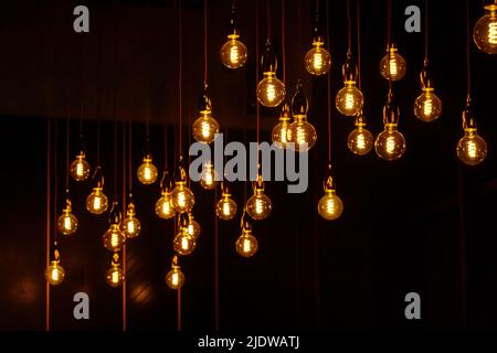 Many vintage light bulbs hanging on cords and glowing against black background at night Stock Photo