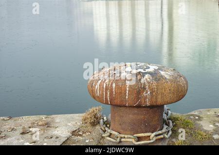 A bollard port by a harbour. Rusty mooring bollard cast iron at pier shore. Sky and water background copyspace. Securing anchor point to prevent Stock Photo