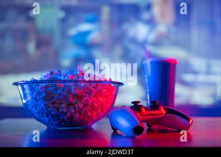 Cozy home relaxation near the TV - popcorn in a transparent glass bowl, soda drink in a plastic glass, gamepad on a sports background. An American foo Stock Photo