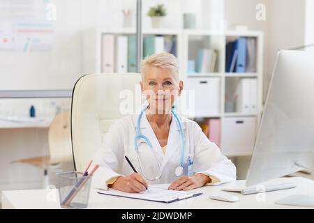 Portrait of serious female doctor in lab coat wearing stethoscope around neck sitting at table in office and filling documents Stock Photo