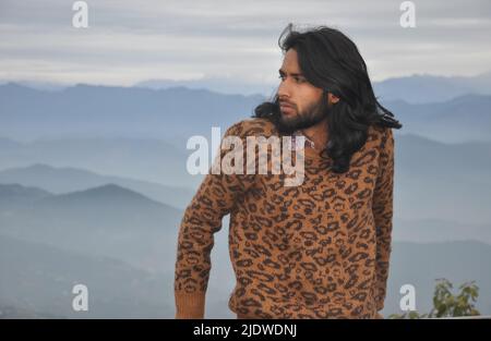 A long haired and bearded Indian young man looking sideways while standing against the background of mountains Stock Photo