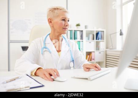 Serious busy mature female doctor in lab coat sitting at desk and using computer while searching for medical researches on internet Stock Photo