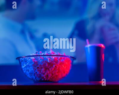 Symbols of a comfortable stay at home near the TV - popcorn in a large glass bowl and a soda drink in a plastic glass with a straw against the backdro Stock Photo