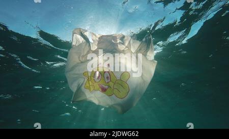 Plastic pollution, used plastic bag with smiley face slowly drifting underwater in sun rays on dawn. Low-angle shot, Backlight (Contre-jour). Plastic Stock Photo