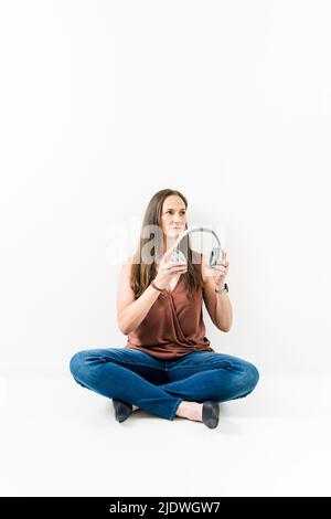 Woman sitting cross legged on ground putting headphones on Stock Photo