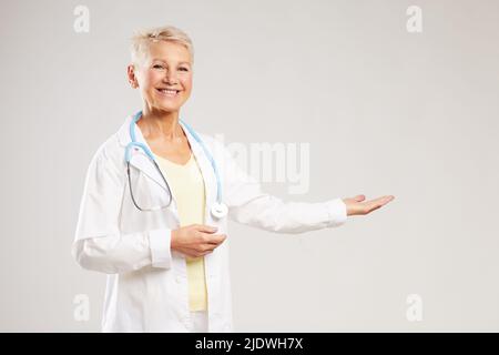 Cheerful excited mature female doctor in white coat standing against blank background and gesturing hands while advertising medical clinic Stock Photo