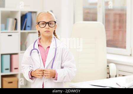 Portrait of serious little girl in big spectacles wearing white coat and stethoscope on neck standing in modern office, she working as doctor Stock Photo