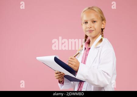 Portrait of content pretty girl in doctors dress standing against pink background and making notes in clipboard Stock Photo