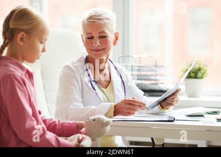 Smiling kind mature female pediatrician with stethoscope sitting at table and making medical history notes while listening to kid at appointment Stock Photo