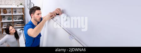 Happy Woman Looking At Electrician Installing Security System Motion Detector On Wall Stock Photo
