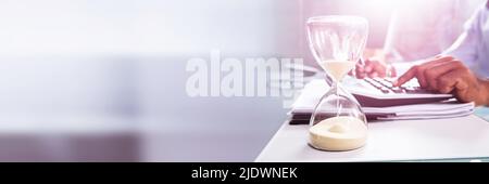 Close-up Of Hourglass In Front Of Businessperson's Hand Calculating Invoice Using Calculator Stock Photo