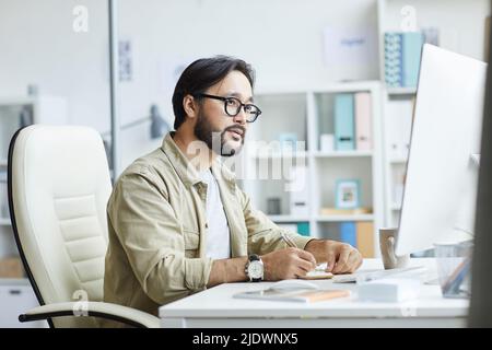 Busy young Asian bearded designer in glasses sitting at desk and using computer while working on project Stock Photo