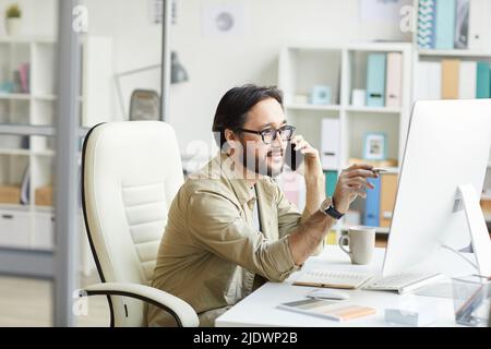 Smart young Asian programmer in casual shirt sitting at table with notepads and pointing at computer monitor while talking to customer by phone Stock Photo