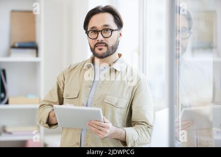 Portrait of serious young Asian java developer with beard standing in office and using tablet as planner Stock Photo