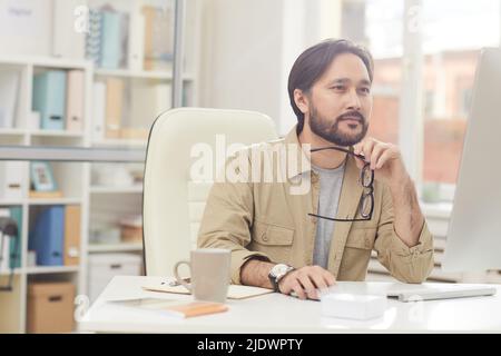 Serious thoughtful young Asian programmer sitting at table and holding eyeglasses while creating design solution Stock Photo