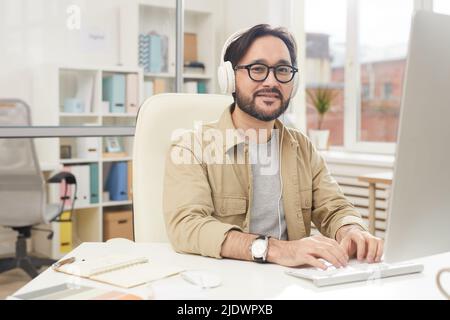 Content handsome young Asian designer in headphones sitting at table and typing on keyboard while enjoying music during work Stock Photo