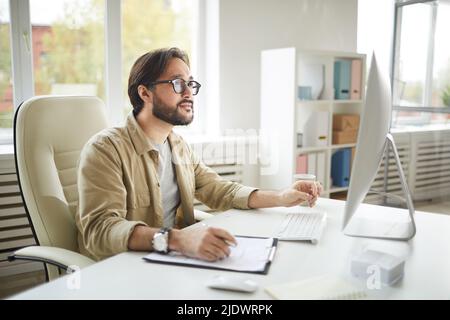 Serious handsome young Asian IT-engineer in casual shirt sitting at table in office and making notes in paper while using computer Stock Photo