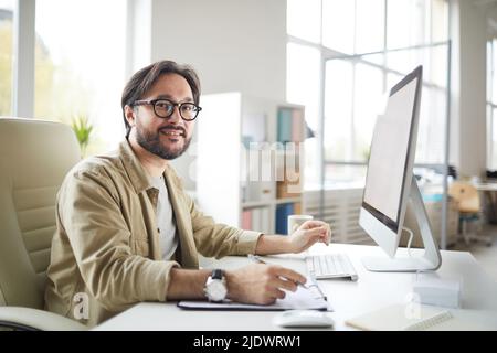 Portrait of cheerful handsome young Asian software engineer in khaki shirt sitting at desk and using modern computer Stock Photo