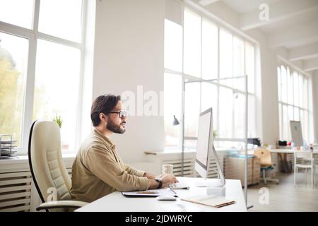 Serious busy young Asian coder in casual shirt and glasses sitting at table and using computer while working with computer language Stock Photo