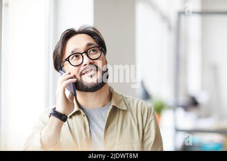 Positive pensive young Asian man with beard sitting by window and using mobile phone for effectively communication Stock Photo