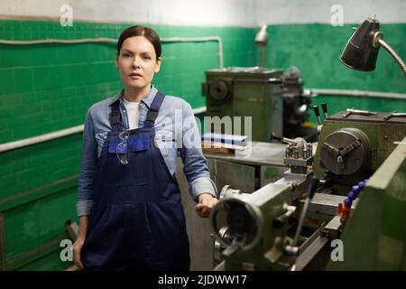 Portrait of serious middle-aged woman in overall with hanging safety goggles standing at manual lathe in industry workshop Stock Photo