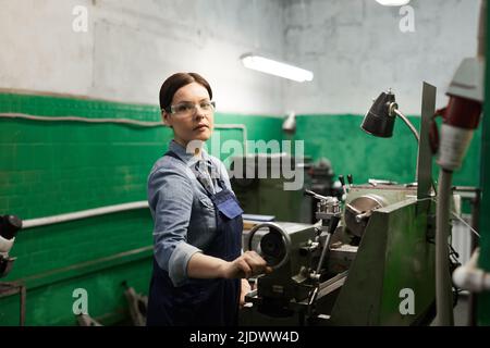 Portrait of confident attractive female engineer in safety goggles using manual lathe while working with metal pieces Stock Photo