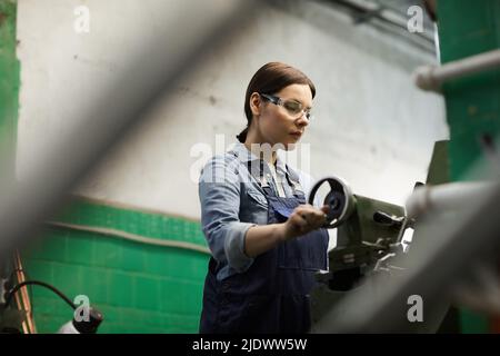 Serious middle-aged female factory worker in protective goggles using tailstock while processing detail on manual lathe Stock Photo